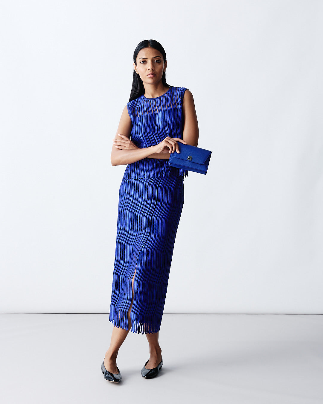 Detailed product shot of a cobalt blue sleeveless top and matching maxi skirt with a textured, ribbed fringe design. The outfit is paired with a coordinated blue envelope clutch bag.