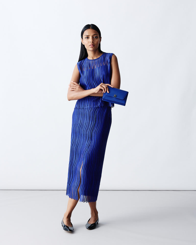 Detailed product shot of a cobalt blue sleeveless top and matching maxi skirt with a textured, ribbed fringe design. The outfit is paired with a coordinated blue envelope clutch bag.