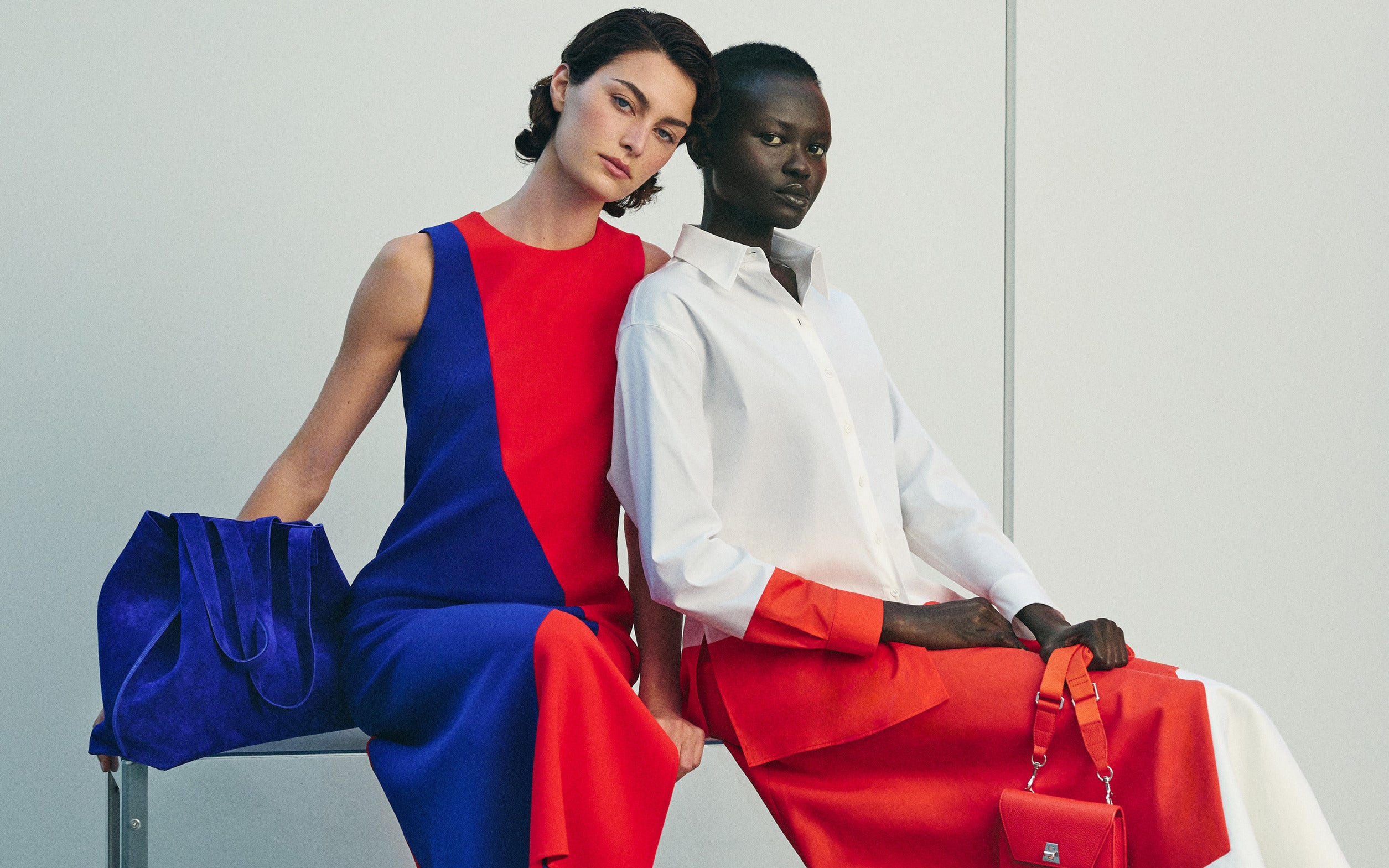 Detailed product shot of a sleeveless red and blue geometric shift dress paired with a vibrant blue suede tote bag. A matching crisp white button-down cotton shirt with orange details is also featured.