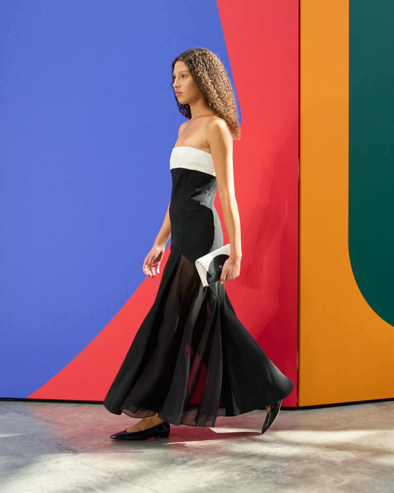 A model walks in profile wearing a strapless floor-length gown with a white bandeau bodice and a black trumpet skirt with sheer chiffon hem. She carries a black and white clutch and wears black flats, against a bold multicolour geometric backdrop