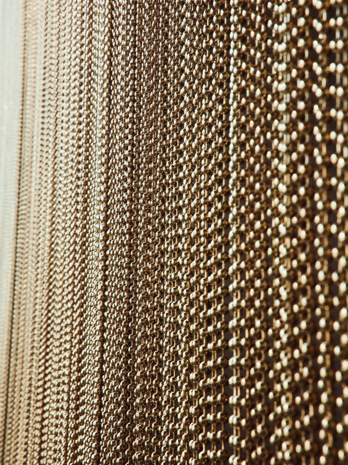 A close-up detail of a metallic beaded curtain, showing the intricate texture of individual golden spheres linked into shimmering vertical chains.