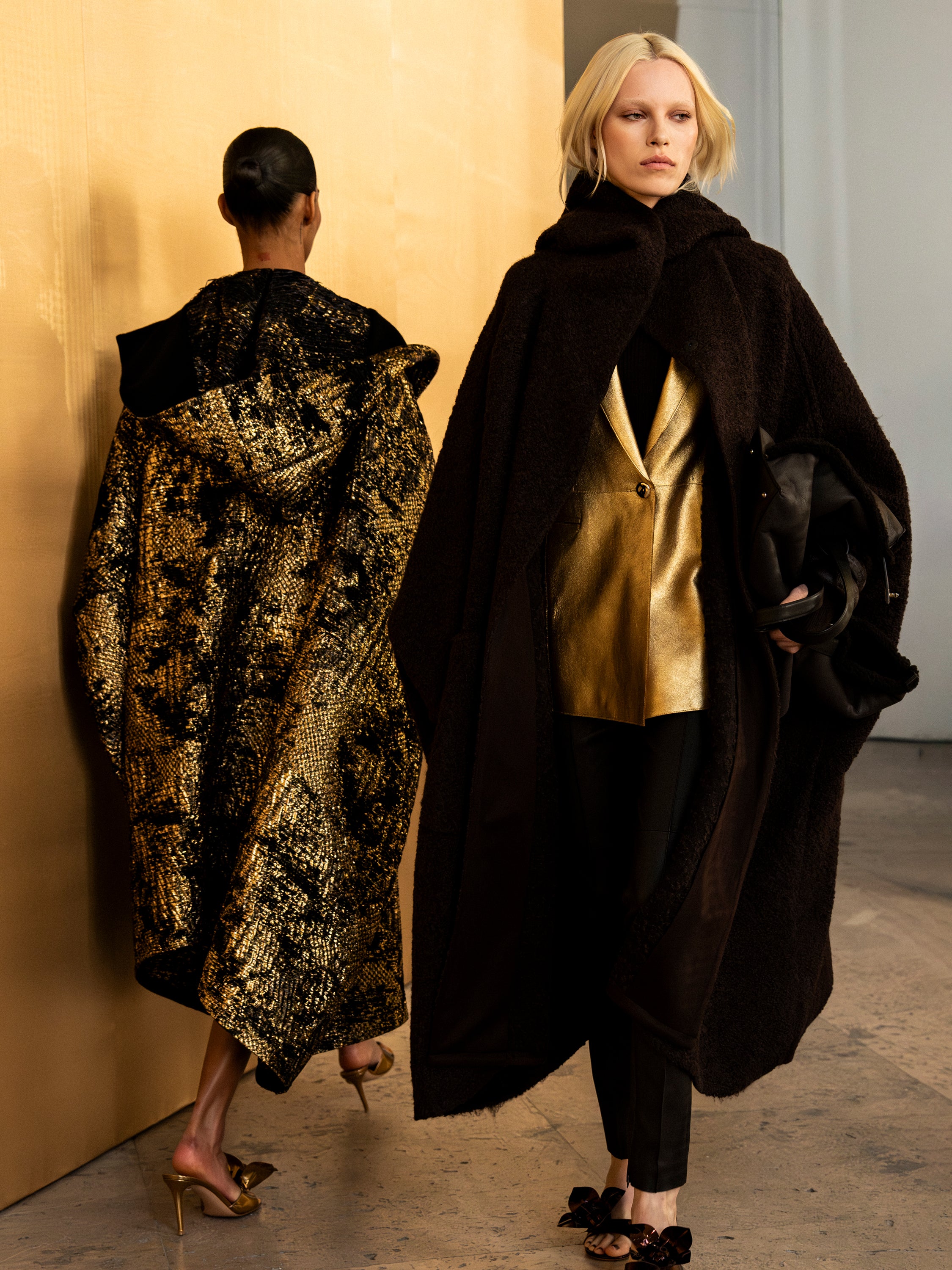 Two models showcase luxurious textures, featuring a voluminous dark textured coat over a metallic gold blazer and a shimmering gold-patterned hooded cape.