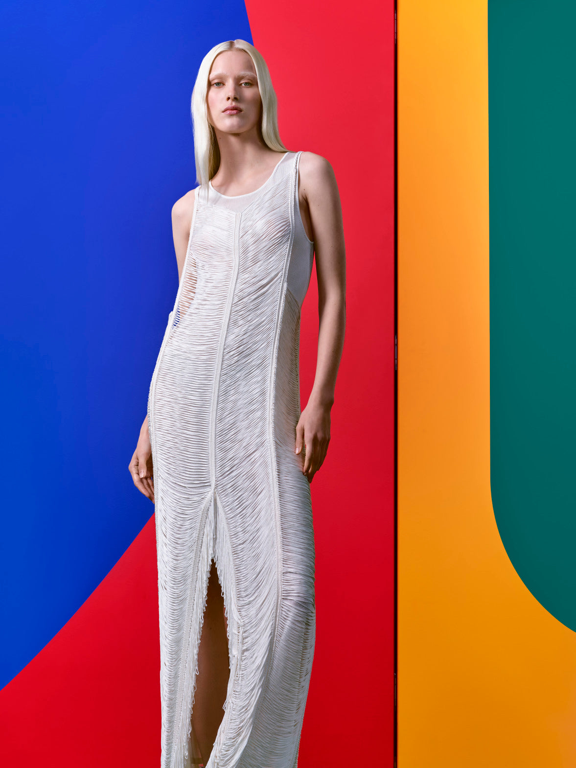 A model wears the Akris Drop Stitch dress in ivory — a floor-length sleeveless gown crafted from an ultra-light silk knit with curved, draped fringe inspired by the contour lines of artist Leon Polk Smith. The layered fringe falls fluidly from the body, styled against a bold split red and blue background