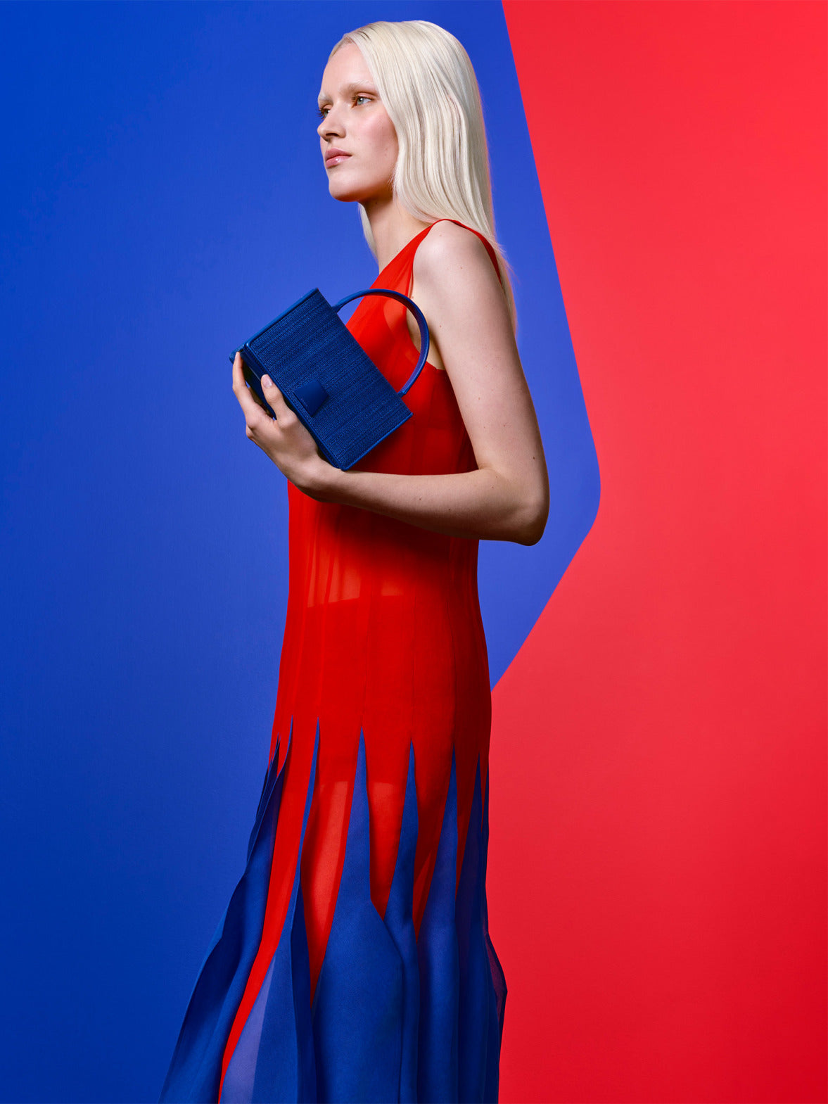 Minimalist geometric fashion shot: A detailed view of a sleeveless red dress with integrated blue flame-like sheer panels, held by a model with a textured blue square handle handbag, set against a split blue and red background.
