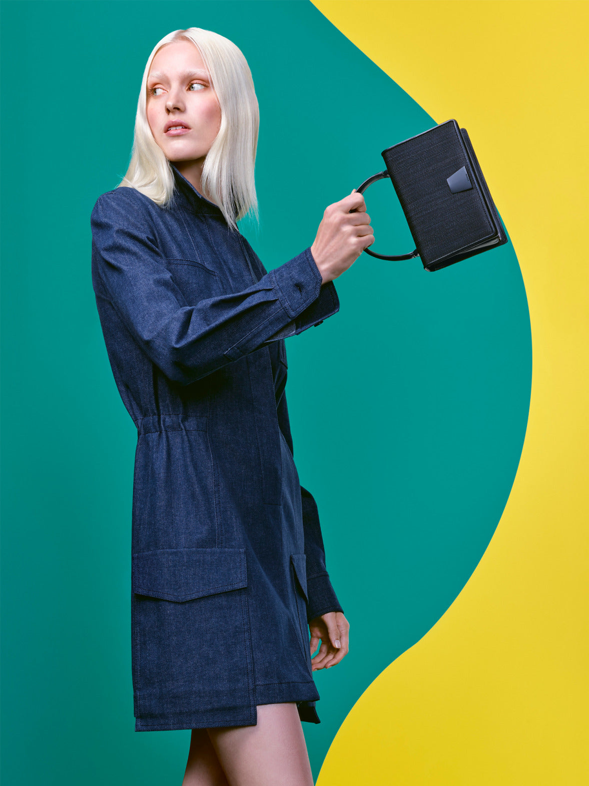 A platinum blonde model wearing a dark denim coat dress, holding a structured black handbag.
The background features a bold, minimalist color-block design in green and yellow.