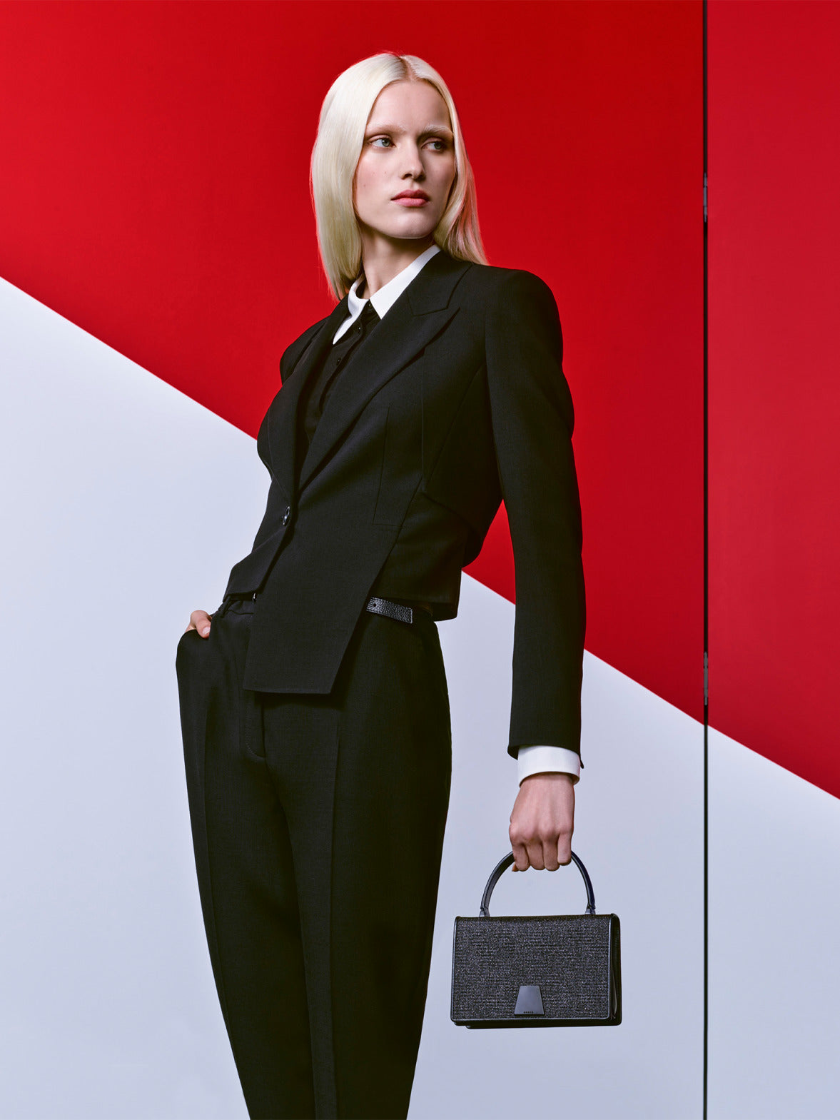 A model with platinum blonde hair in a sharp black suit and tie, posing against a minimalist red and white diagonal background. They are looking to the side while holding a black handbag.