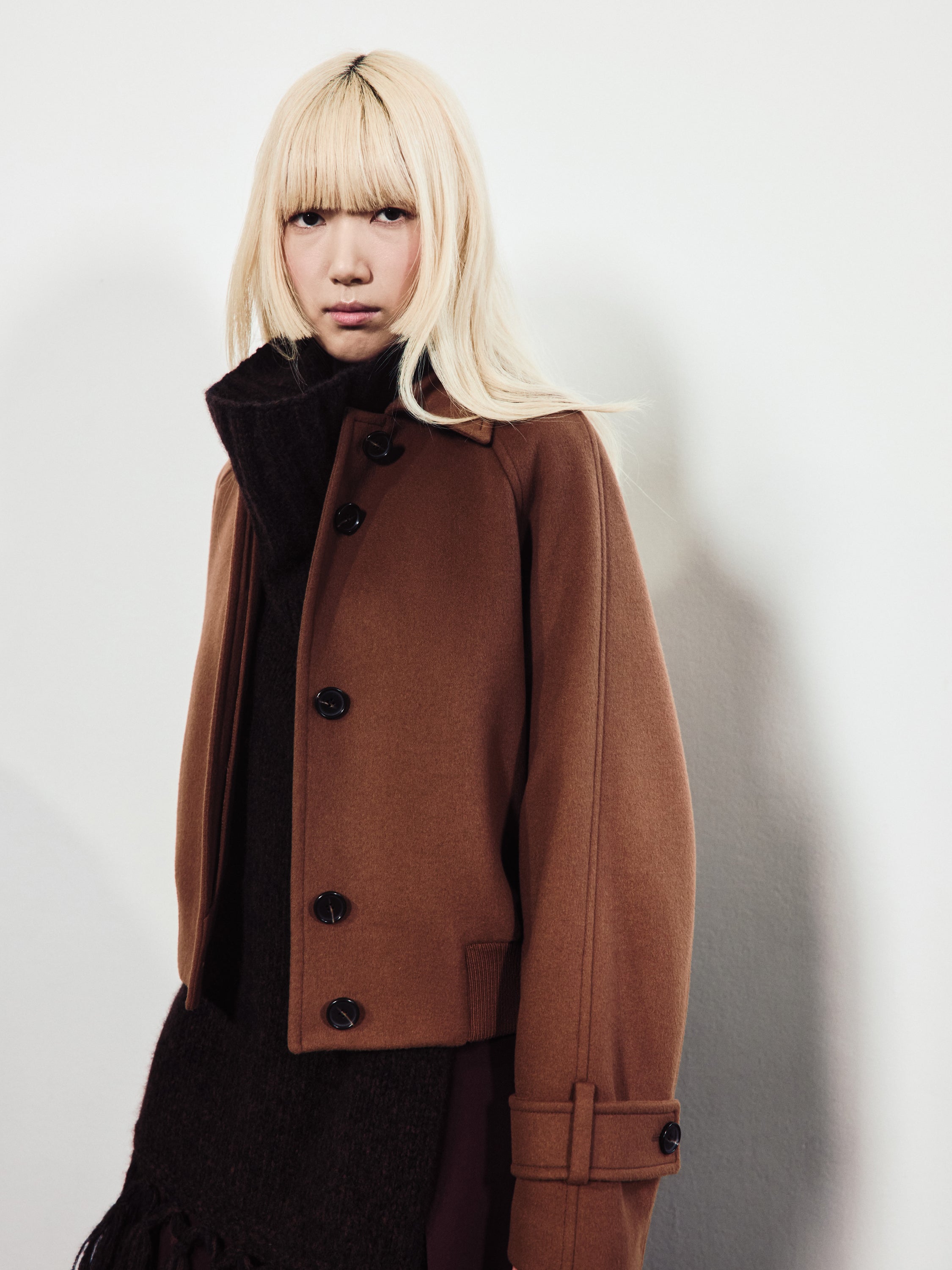 A blonde model wears a cropped, tobacco-brown wool jacket with large black buttons over a thick dark turtleneck sweater.