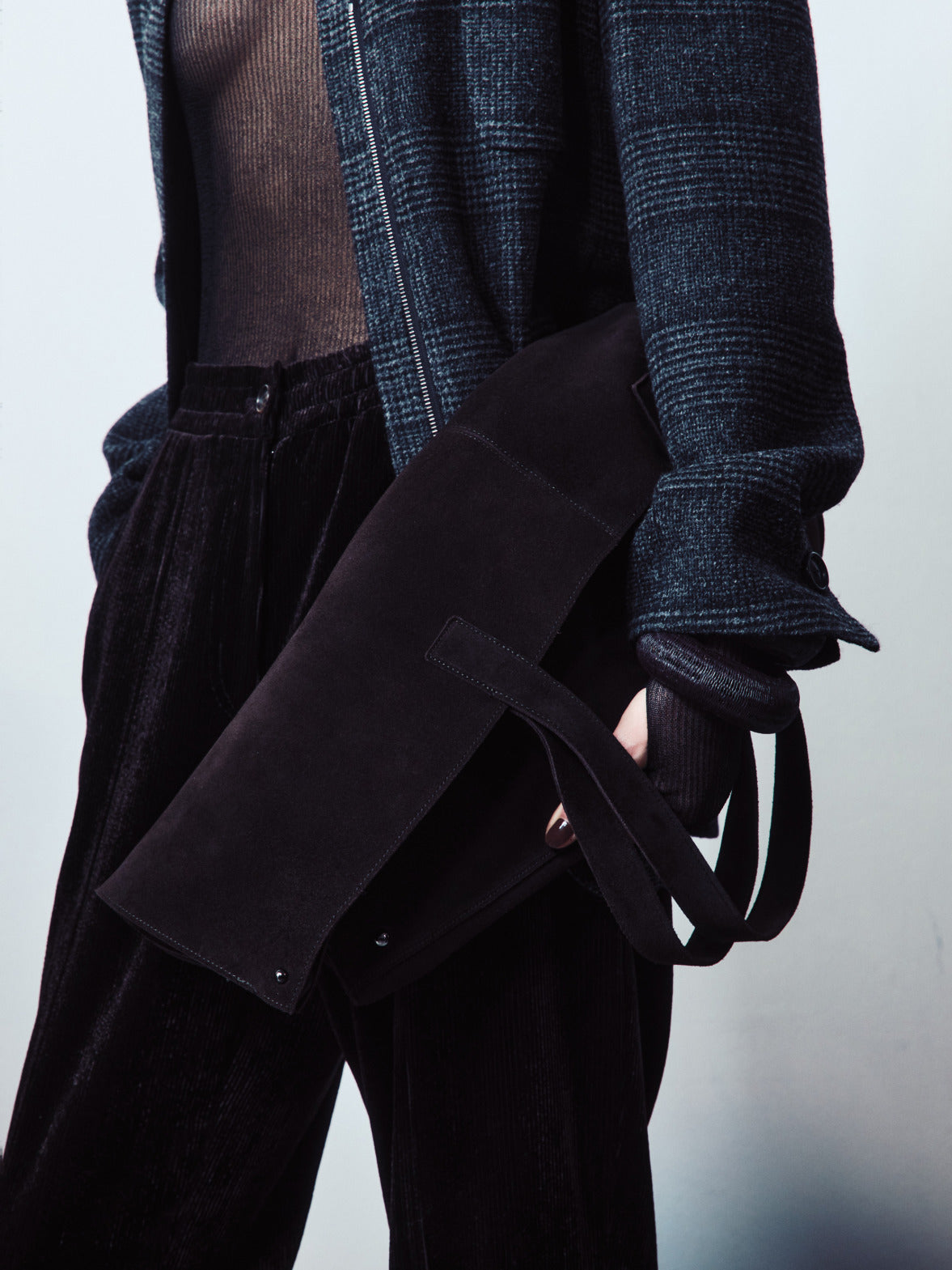 A close-up featuring a large black suede clutch bag paired with a dark teal plaid jacket and textured velvet trousers.