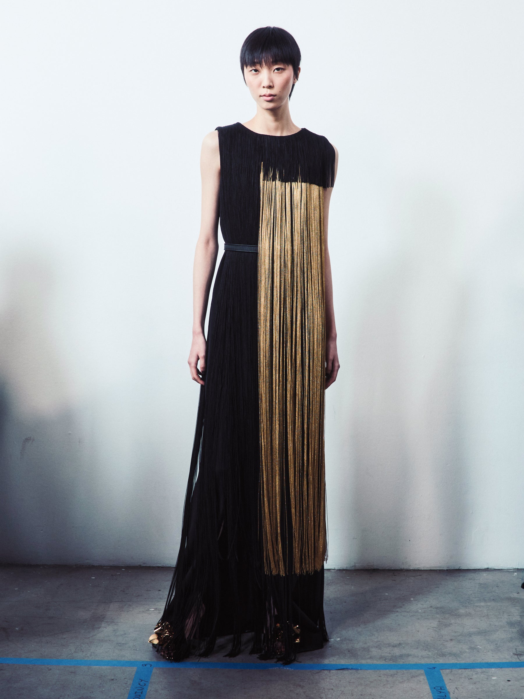 A model wears a sophisticated black sleeveless gown featuring long, floor-length vertical fringes with a striking asymmetrical gold panel.
