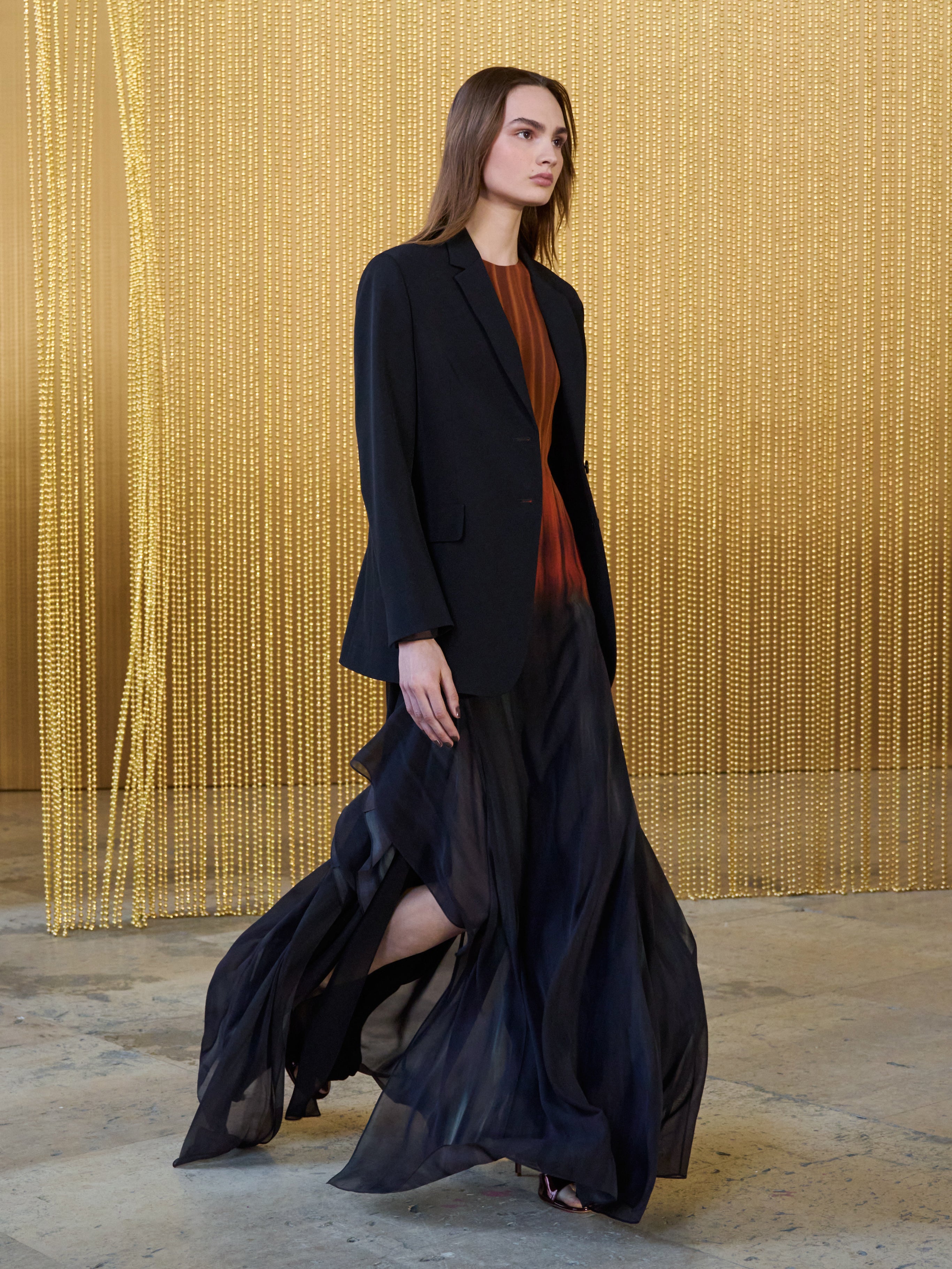 A model walks in a dark blazer over a flowing gown with a red-to-black gradient, set against a shimmering gold beaded curtain.