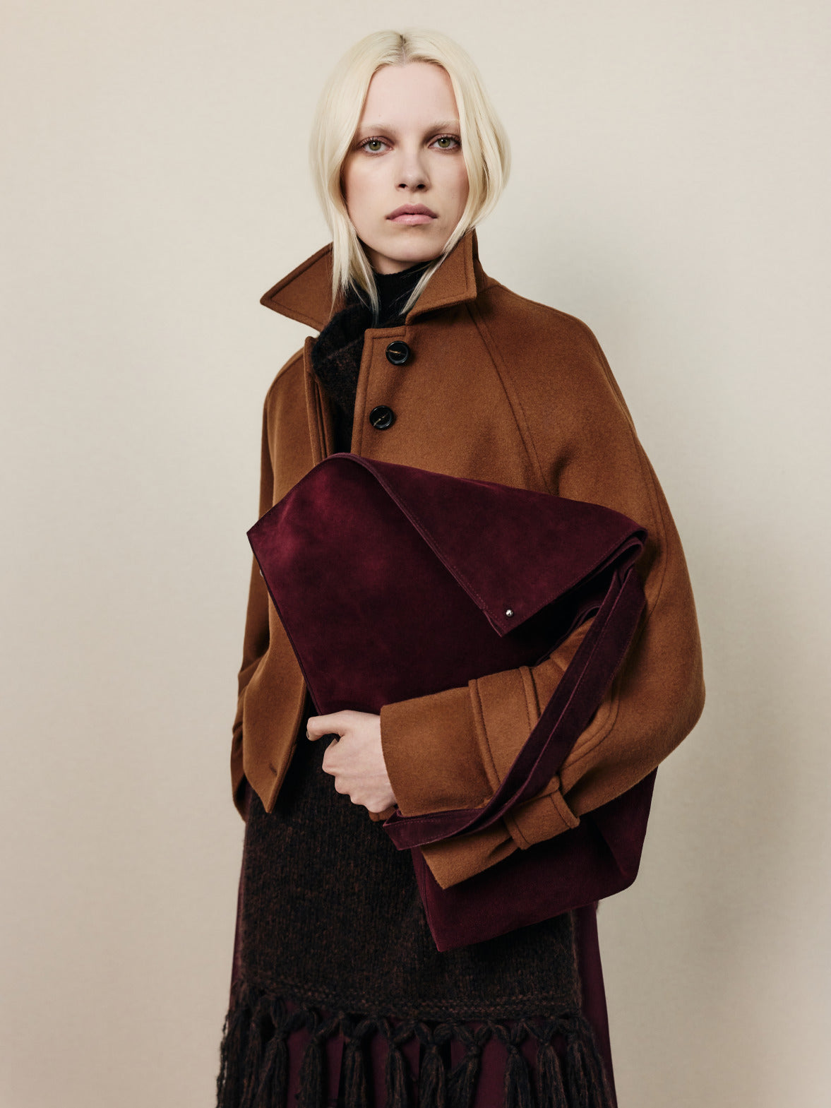Look from the Akris Fall 2026 collection featuring a camel outerwear silhouette and an oversized suede bag in rich burgundy tones.