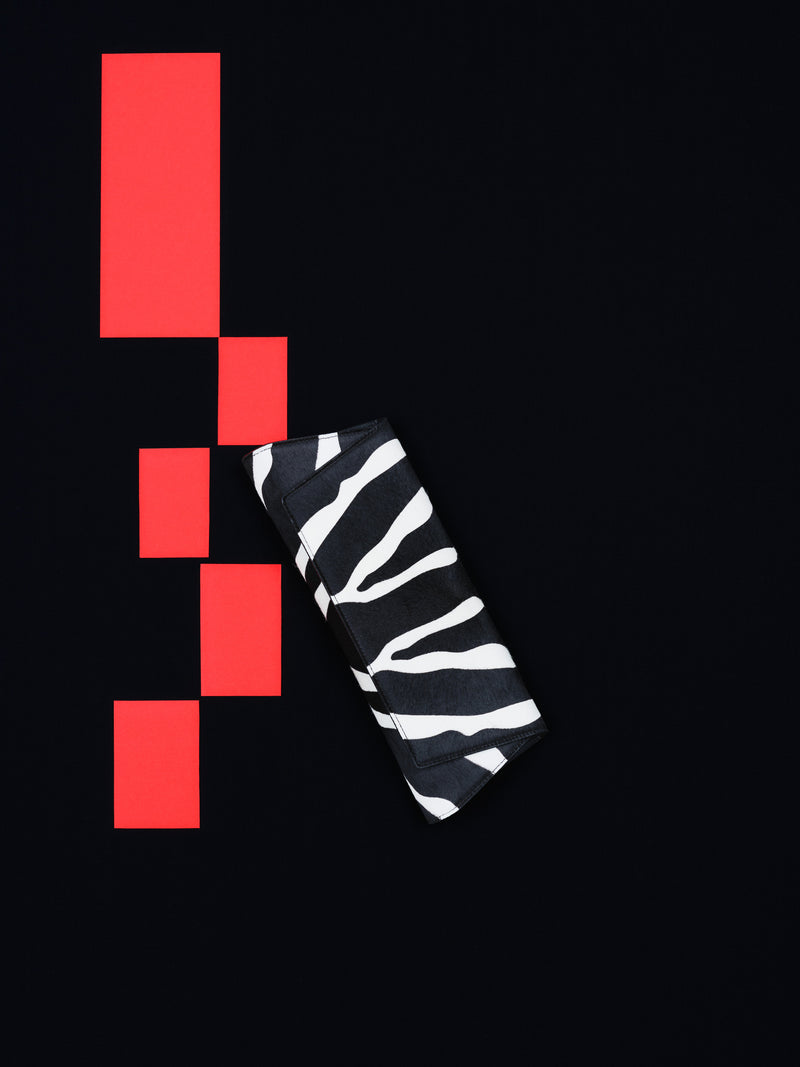 Discover the graphic allure of the Akris Pre-Fall collection, showcasing the elegant Anouk clutch bag in a striking zebra print. Positioned against a minimalist black backdrop with bold red geometric accents, this luxury accessory makes a powerful style statement. Embrace contemporary design and exceptional Swiss craftsmanship.