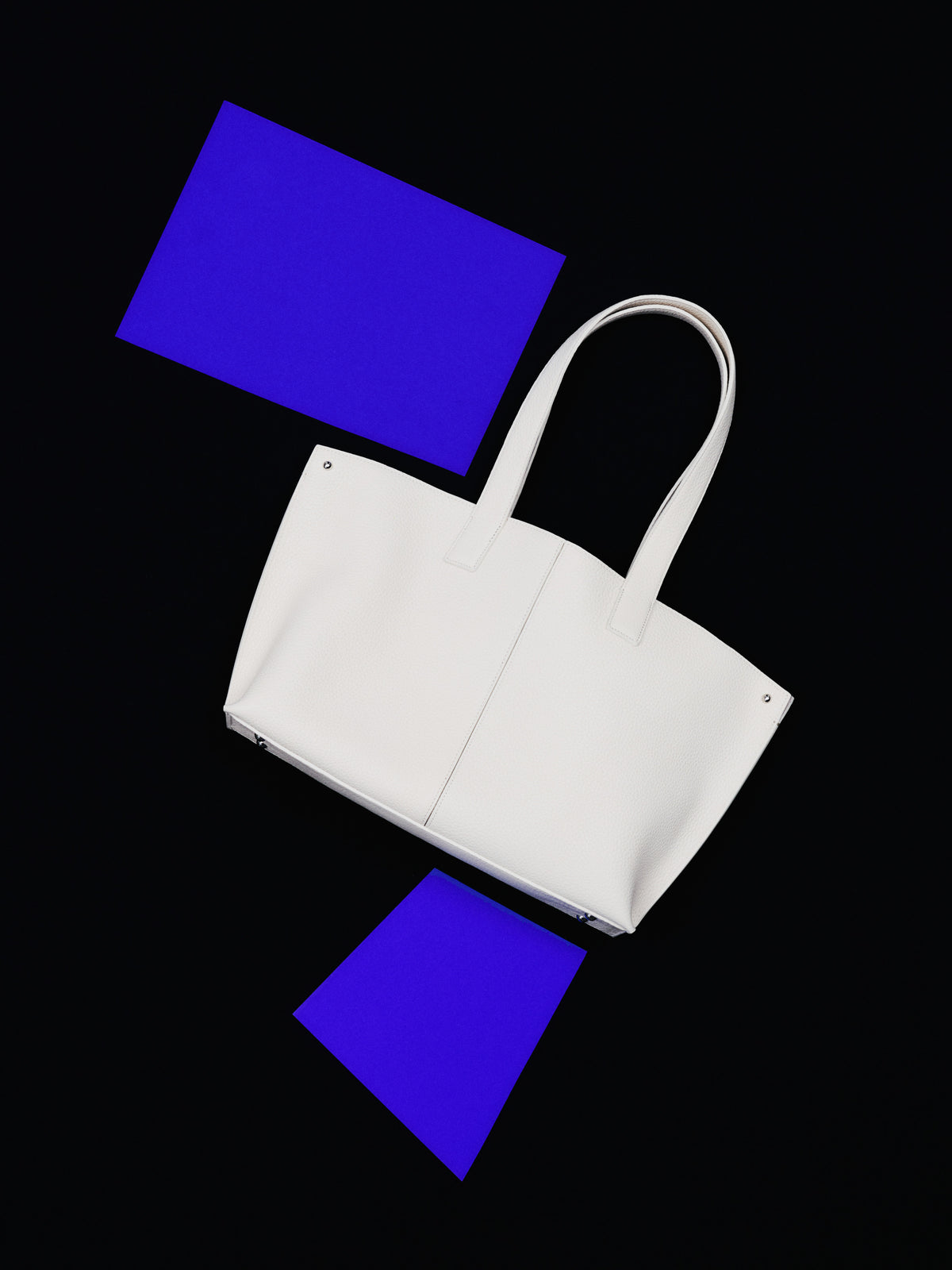 Discover the minimalist perfection of the Akris Pre-Fall collection, highlighting the iconic Ai tote bag in a pristine white leather finish. Artfully arranged against a dramatic black backdrop with vivid electric blue geometric accents, this luxury accessory embodies pure modern elegance. Elevate your collection with timeless Swiss design.