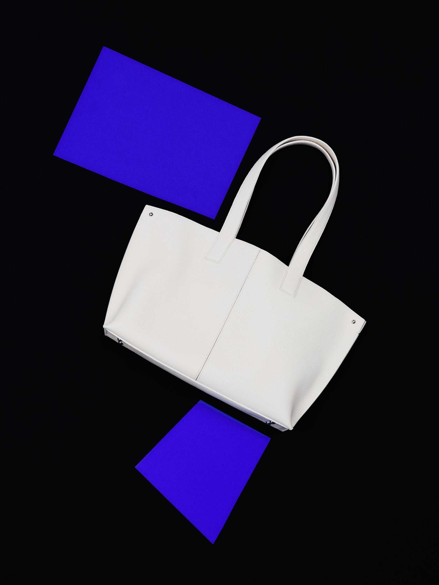 Akris Ai ivory leather shoulder bag with structured handles, shown overhead on a black surface with geometric blue squares, from the new Akris collection.