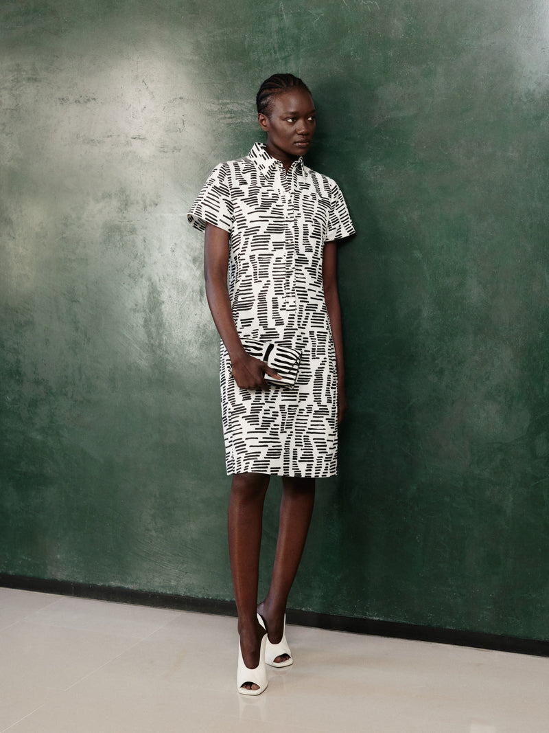 Discover the graphic elegance of the Akris Pre-Fall collection, highlighting a classic shirt dress featuring an abstract black-and-white print. The sophisticated look is paired with a matching luxury clutch bag and white open-toe mules. Elevate your everyday style with effortless modern design and premium Swiss craftsmanship.