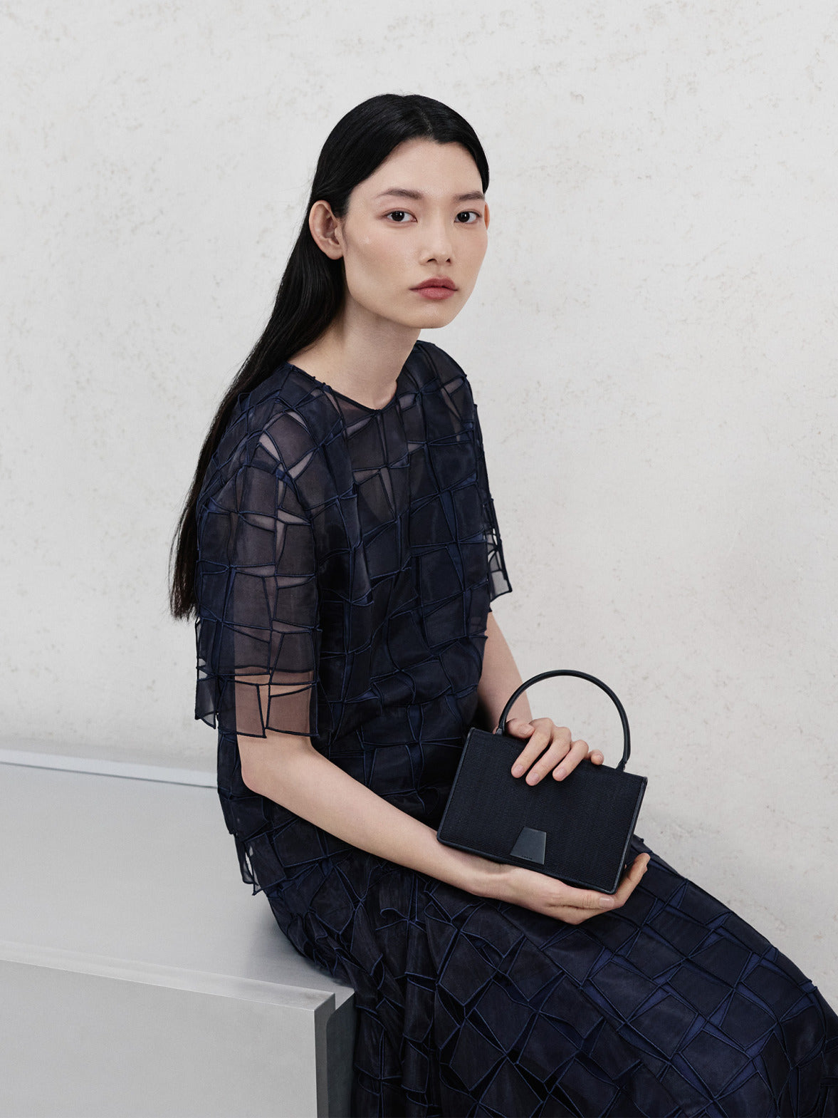 Akris Pre-Fall 2026 collection featuring a dark blue sheer patchwork short-sleeve top and matching skirt. The look is styled with a structured black leather handbag with a top handle, set against a minimalist cream background.