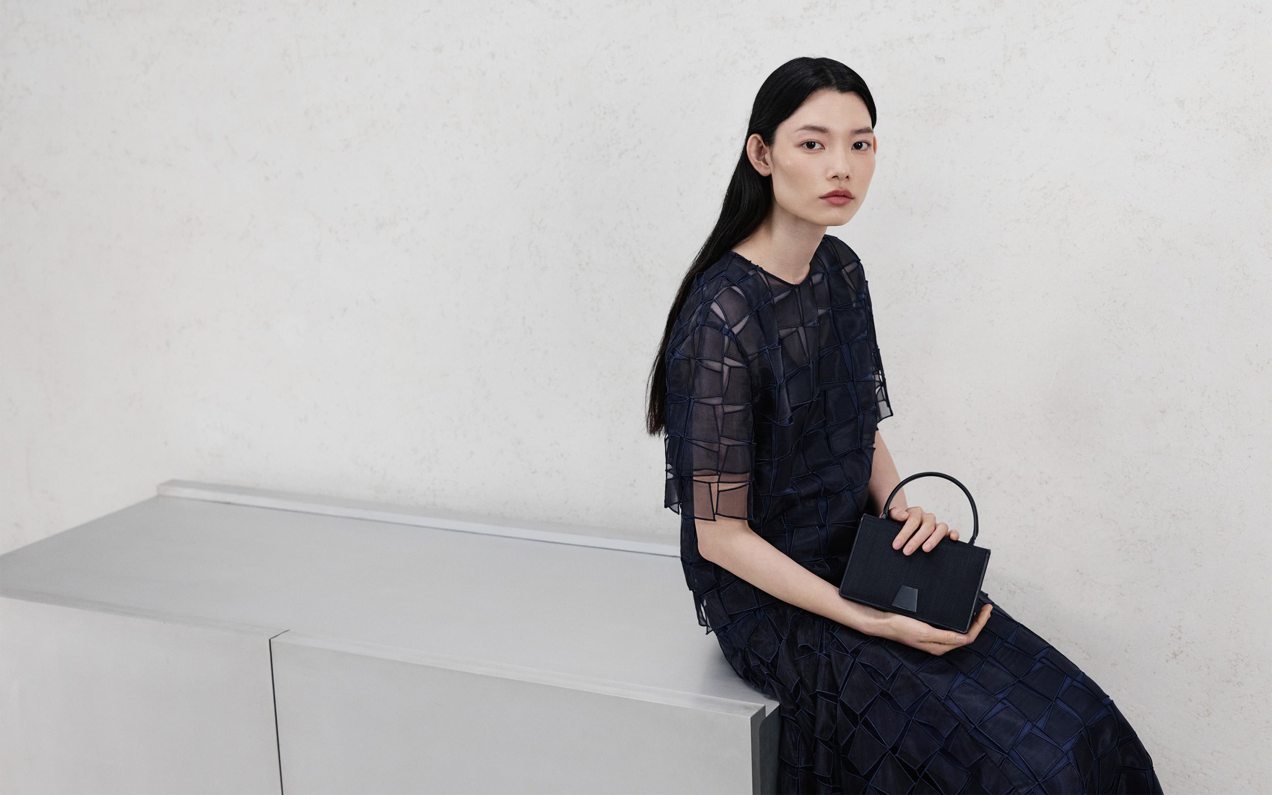 Akris Pre-Fall 2026 collection featuring a dark blue sheer patchwork short-sleeve top and matching skirt. The look is styled with a structured black leather handbag with a top handle, set against a minimalist cream background.