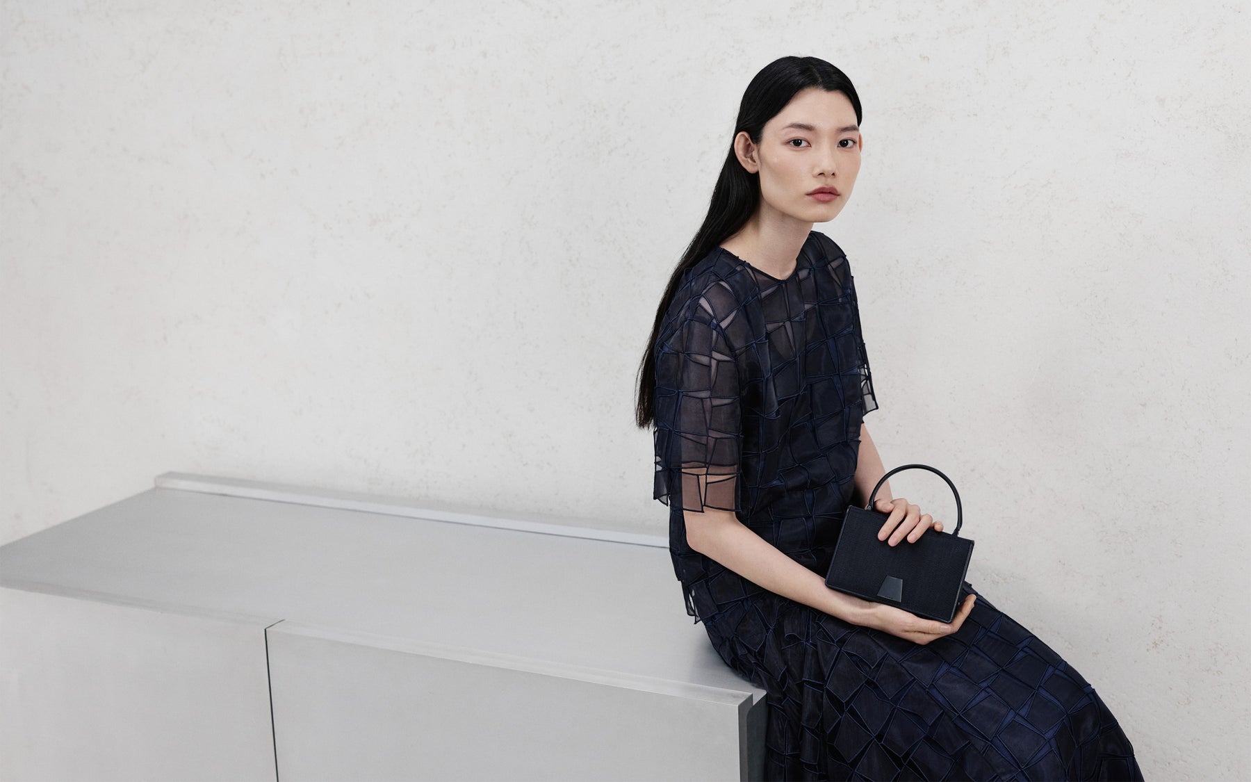Model wearing a navy blue Akris Pre-Fall dress with geometric architectural embroidery, holding a matching denim structural handbag while sitting against a minimalist wall.
