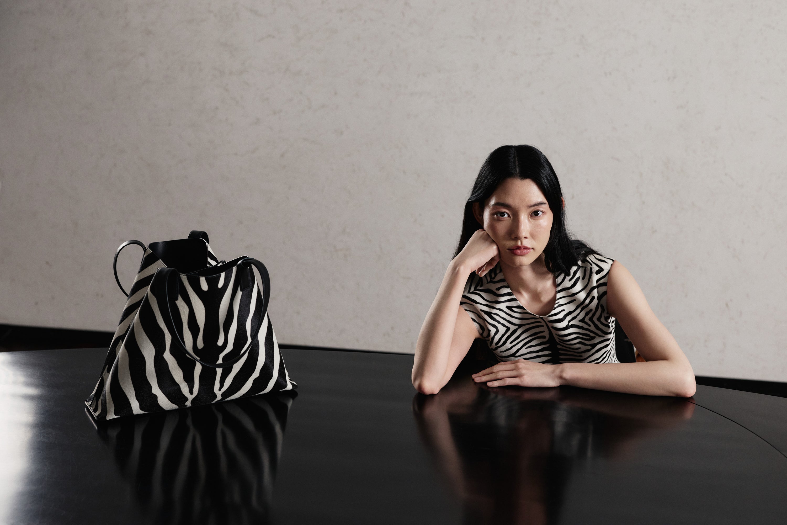Discover the graphic sophistication of the Akris Pre-Fall collection, featuring a timeless sleeveless top in a bold zebra print. Placed elegantly on the table is the iconic Ai tote bag in a matching zebra motif, creating a seamless high-contrast look. Elevate your wardrobe with minimalist shapes and premium Swiss craftsmanship.