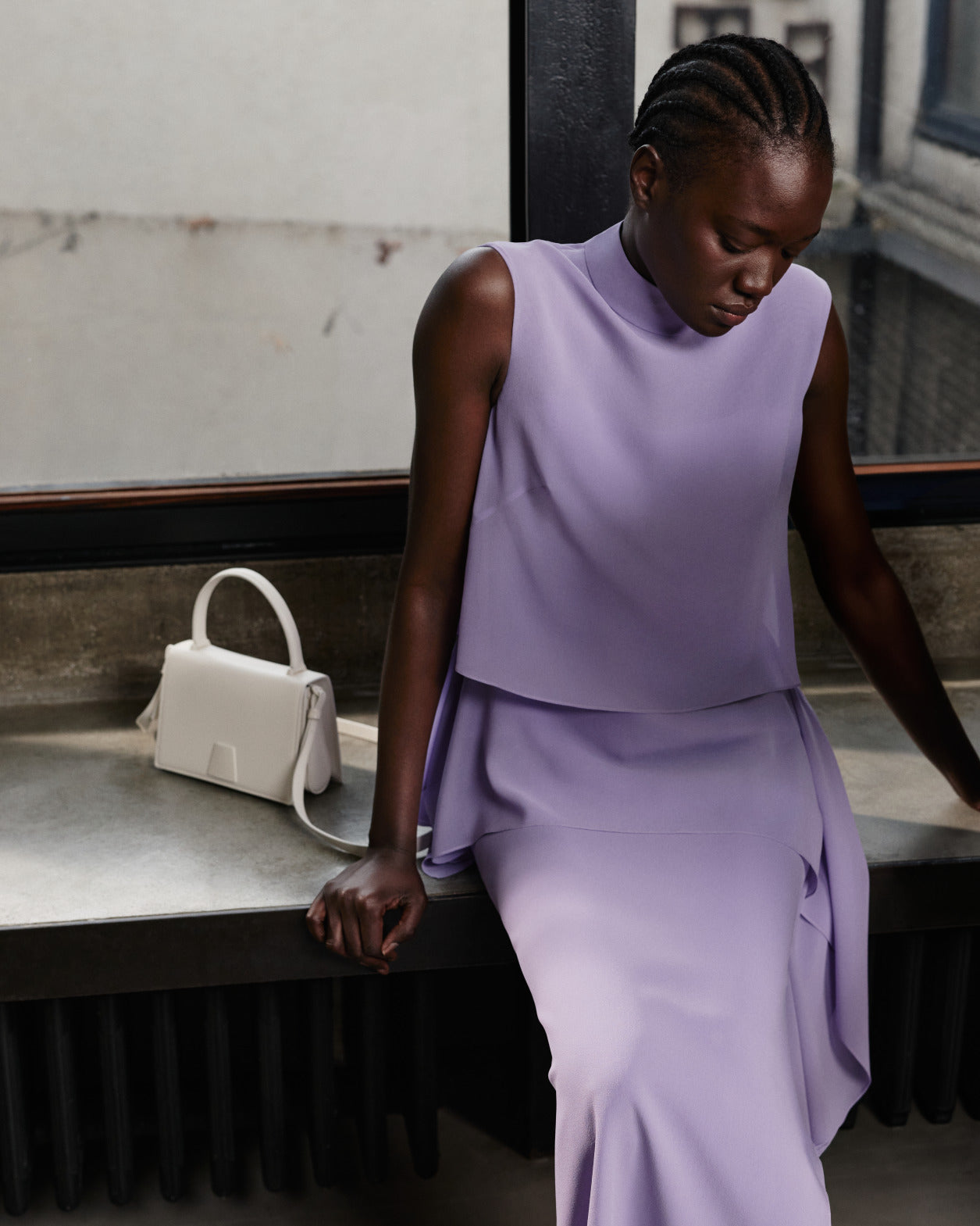 Model wearing a lavender Akris Pre-Fall malva dress , sitting by a window next to a structured white leather shoulder bag.