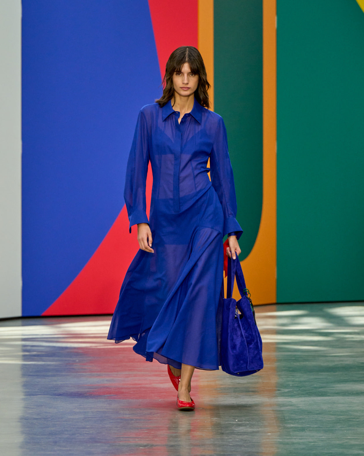 A model walks the runway in a sheer cobalt blue long-sleeve shirt dress, carrying a matching blue suede bag and wearing red flats, against a bold multicolour geometric backdrop.