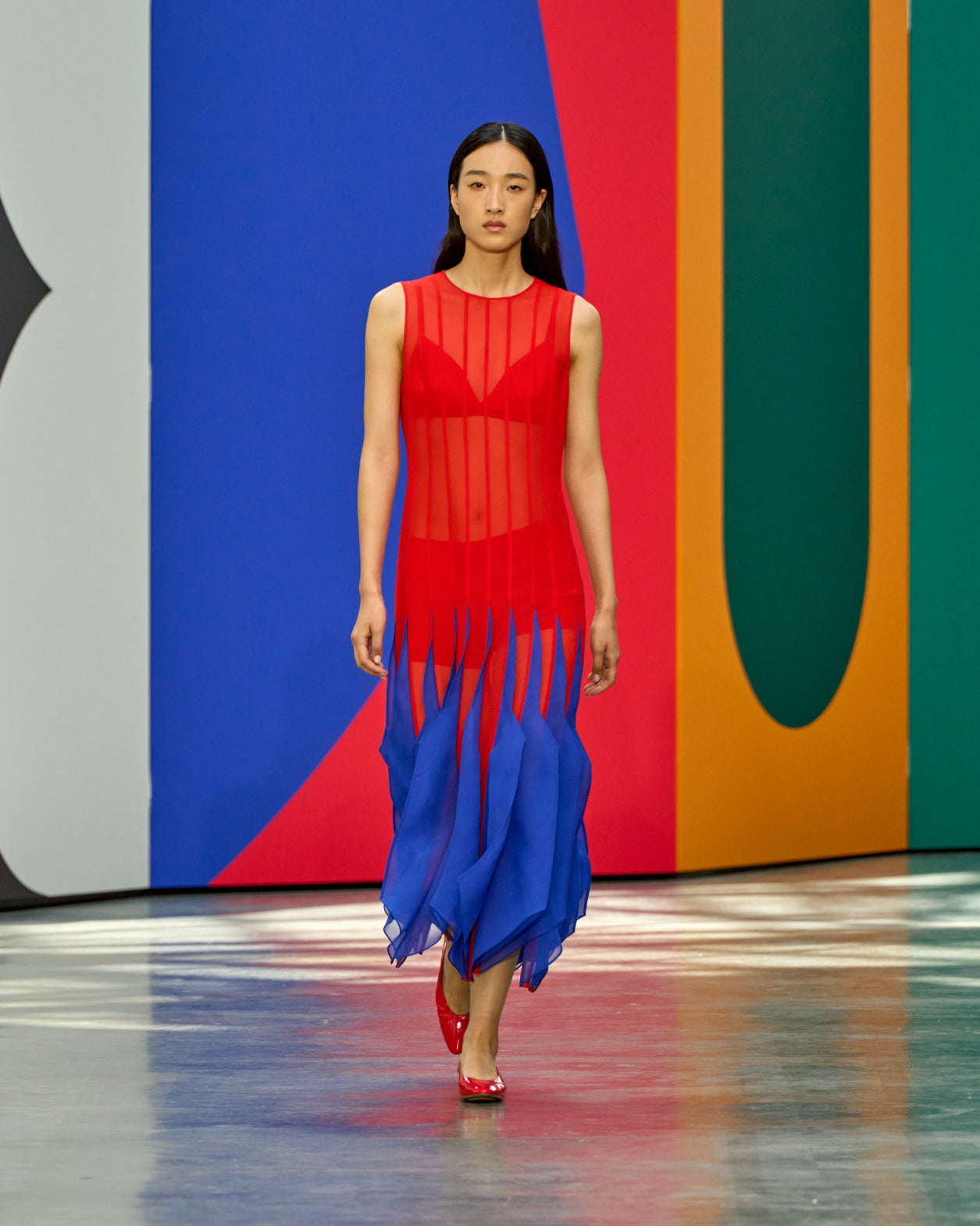 A model walks the runway in a sleeveless red sheer dress with a blue fringe and layered skirt hem, paired with red heels, against a bold multicolour geometric backdrop