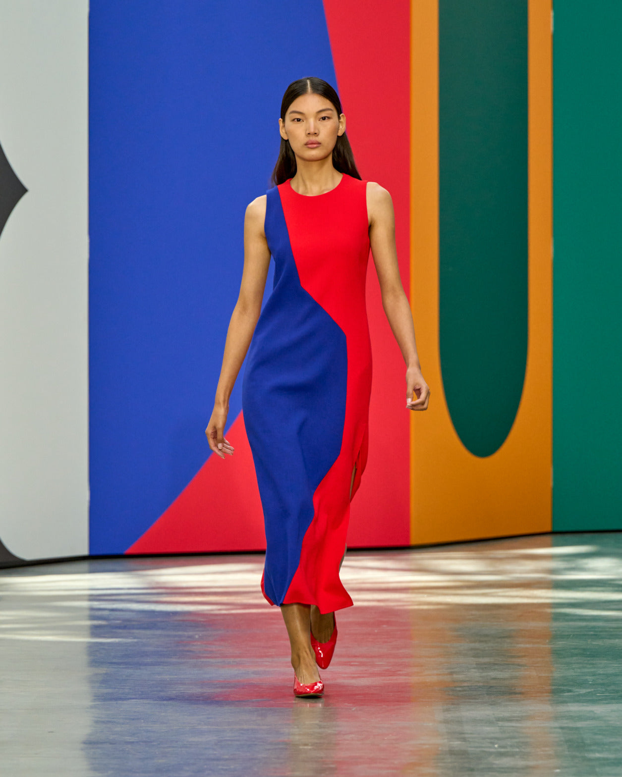 A model walks the runway in the Akris High Plains midi sheath dress in wool double-face, featuring a bold colour-block design in vermillion red and lapis blue with a curved patchwork seam, crew neckline, sleeveless silhouette, and subtle side slit. Paired with red heels against a bold multicolour geometric backdrop.