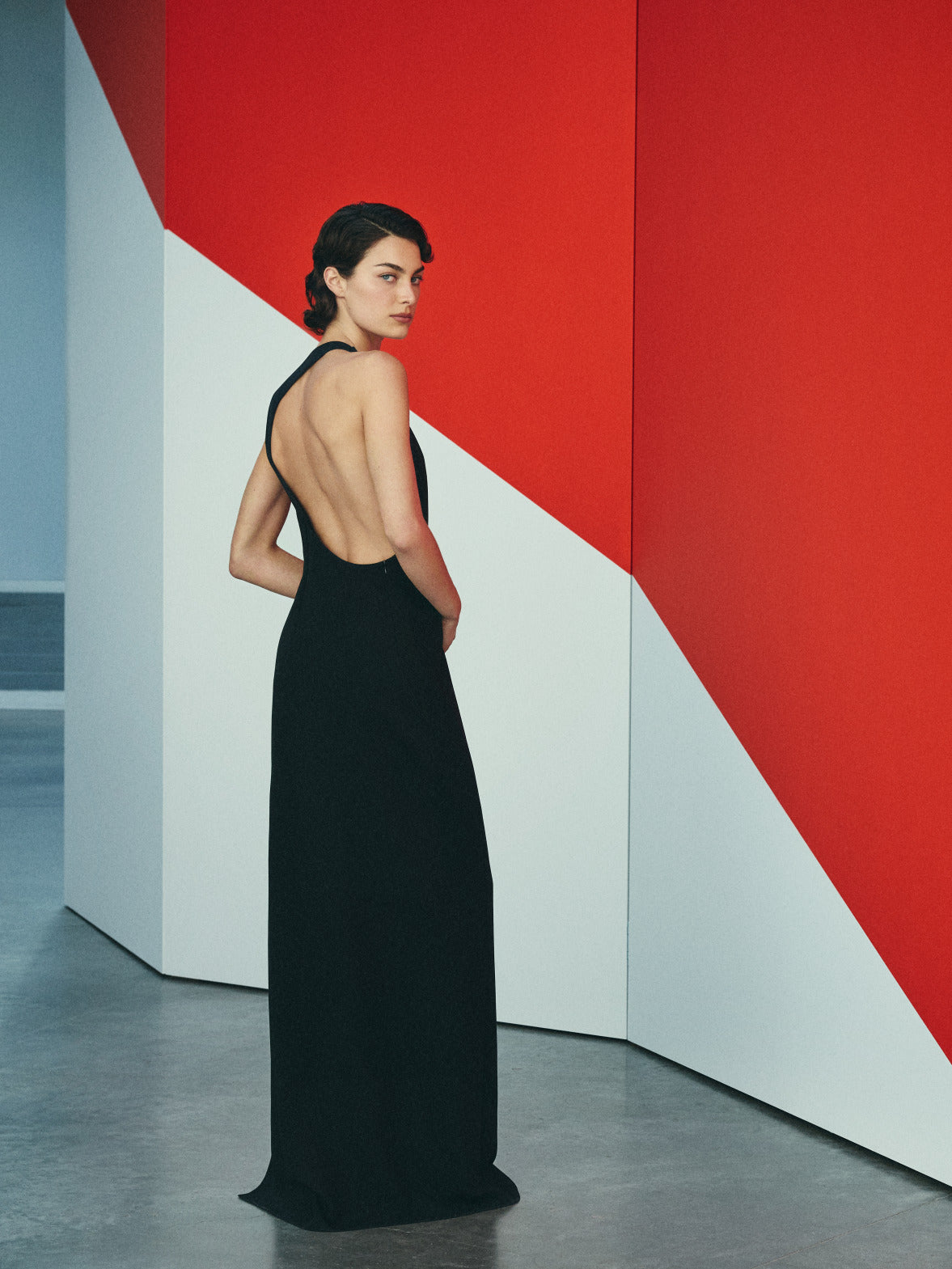 The model wears a floor-length black halter gown with a deep open back. She glances over her shoulder against a minimalist gallery-like backdrop featuring bold geometric shapes in red and white