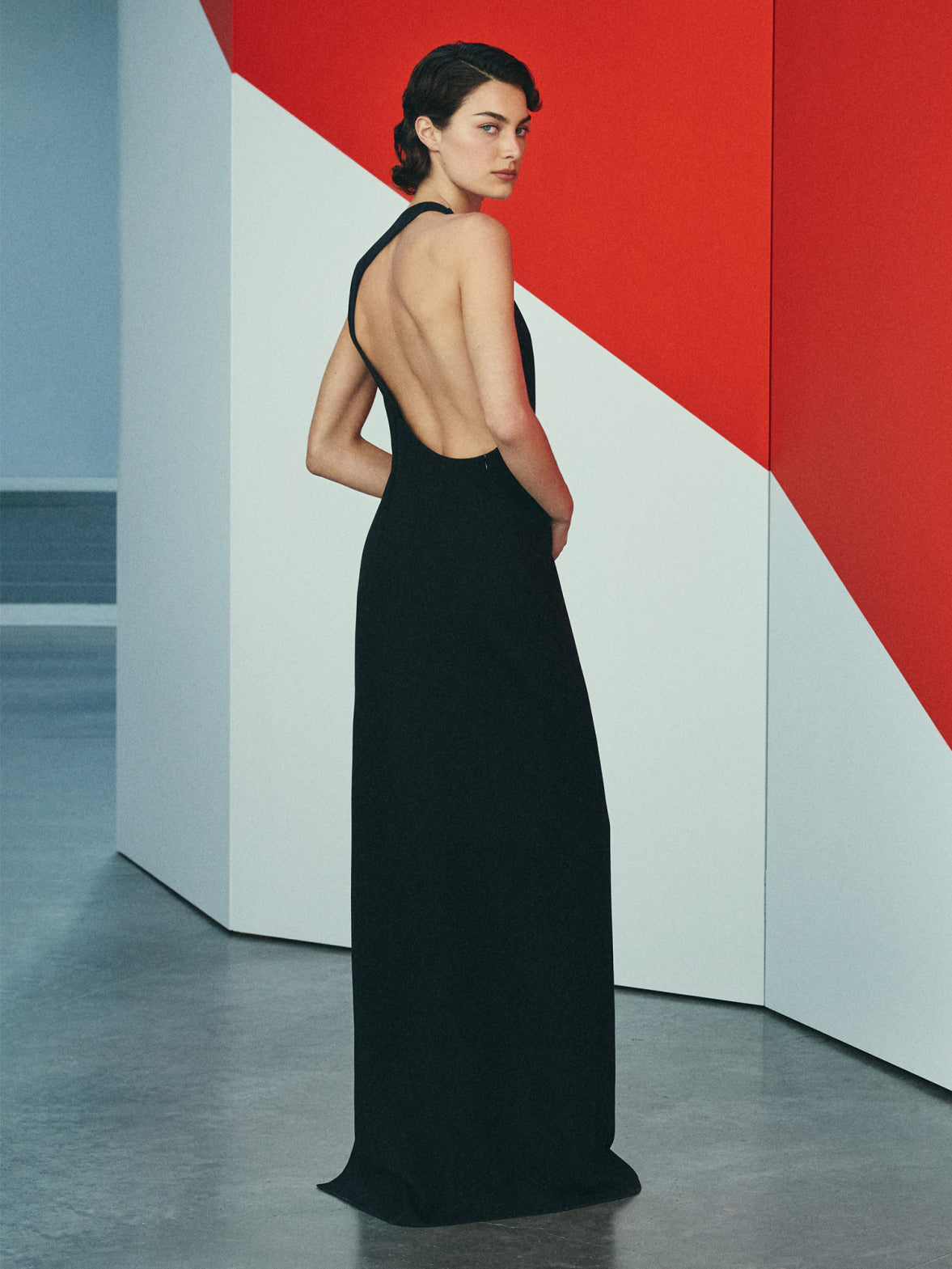 A model in an elegant backless black dress, looking over their shoulder against a geometric red and white background.
The shot emphasizes the minimalist aesthetic and the clean lines of the garment.