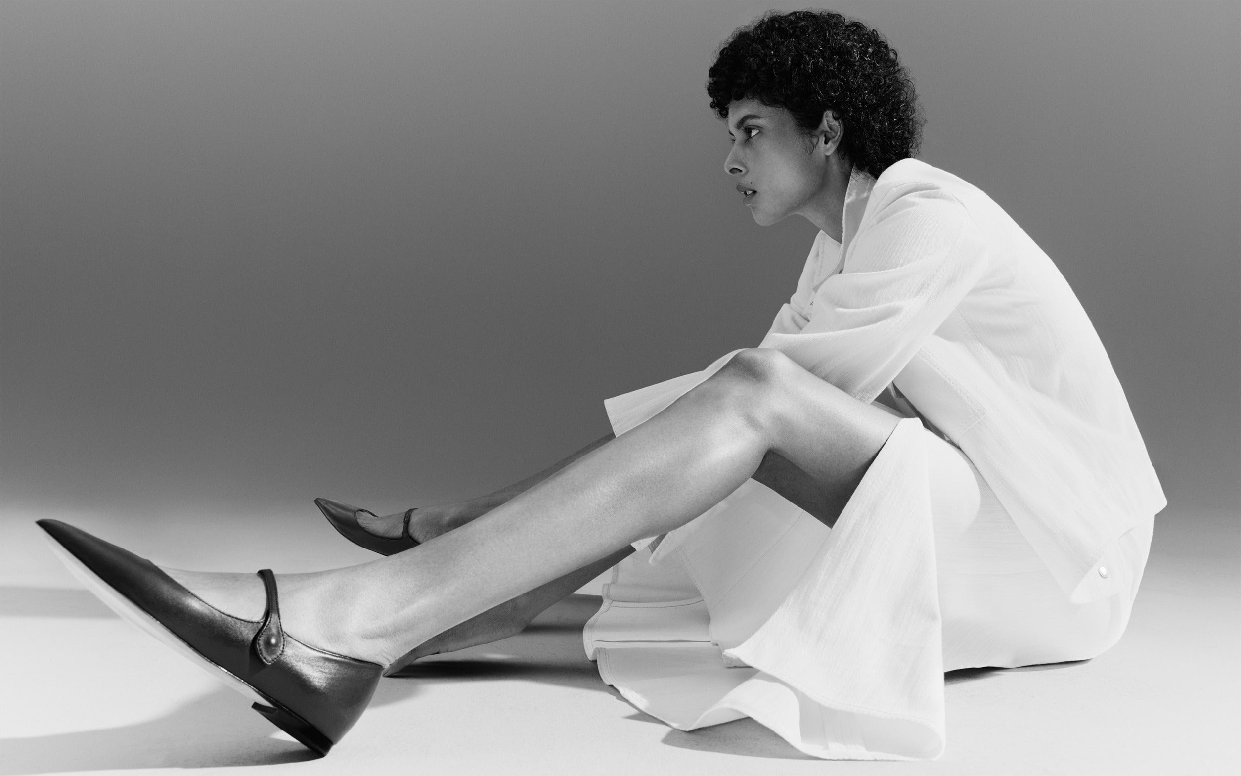 Modernist Akris punto Pre-Fall 2026 editorial look featuring a woman in an all-white textured outfit with a pleated skirt. She wears minimalist black leather pointed-toe Mary Jane flats against a neutral studio backdrop.