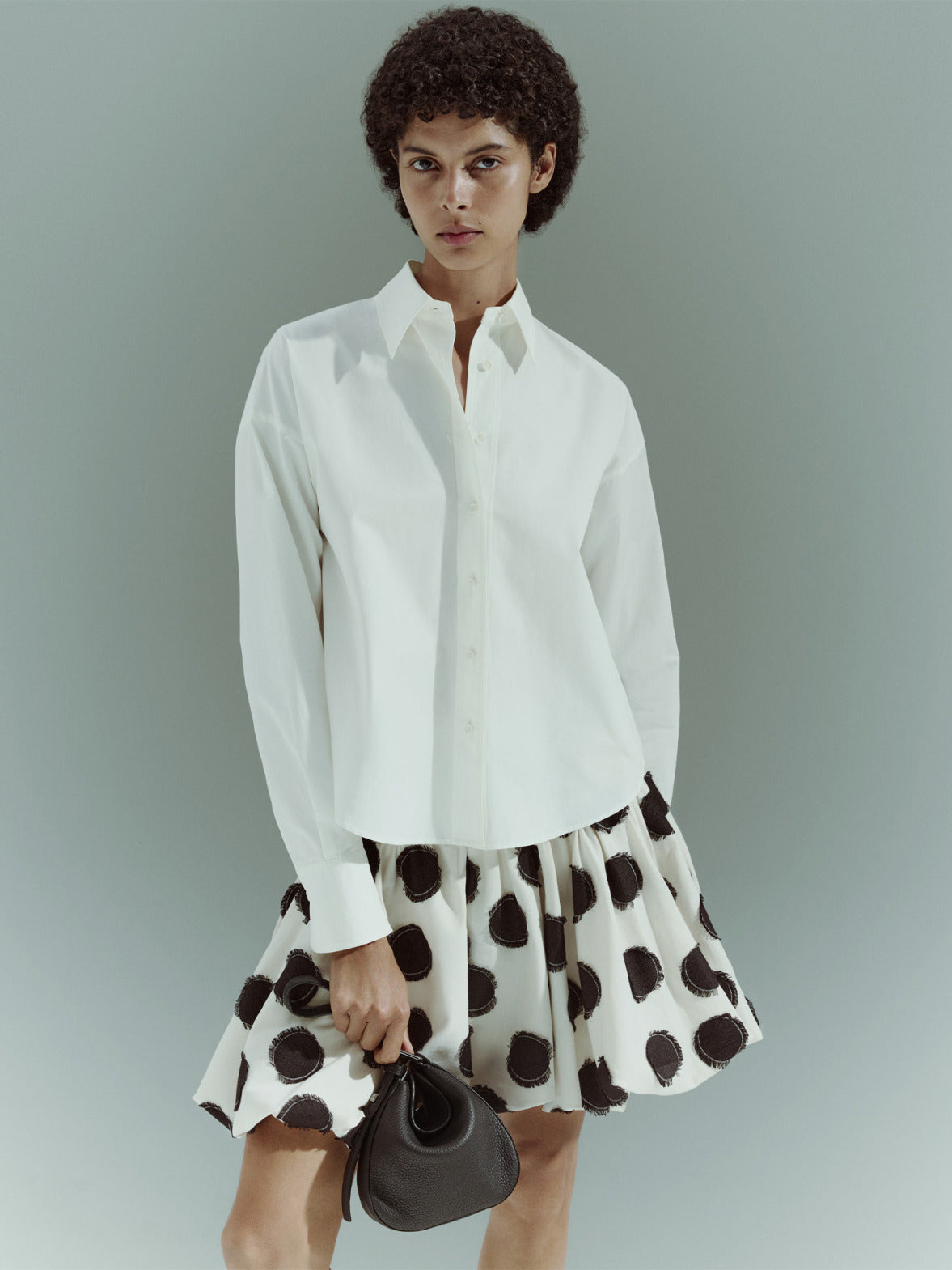 Akris punto Pre-Fall 2026 collection luxury women's apparel.
Features a sleeveless white midi dress with large black polka dots.
Styled next to a classic long-sleeve white button-up cotton shirt.