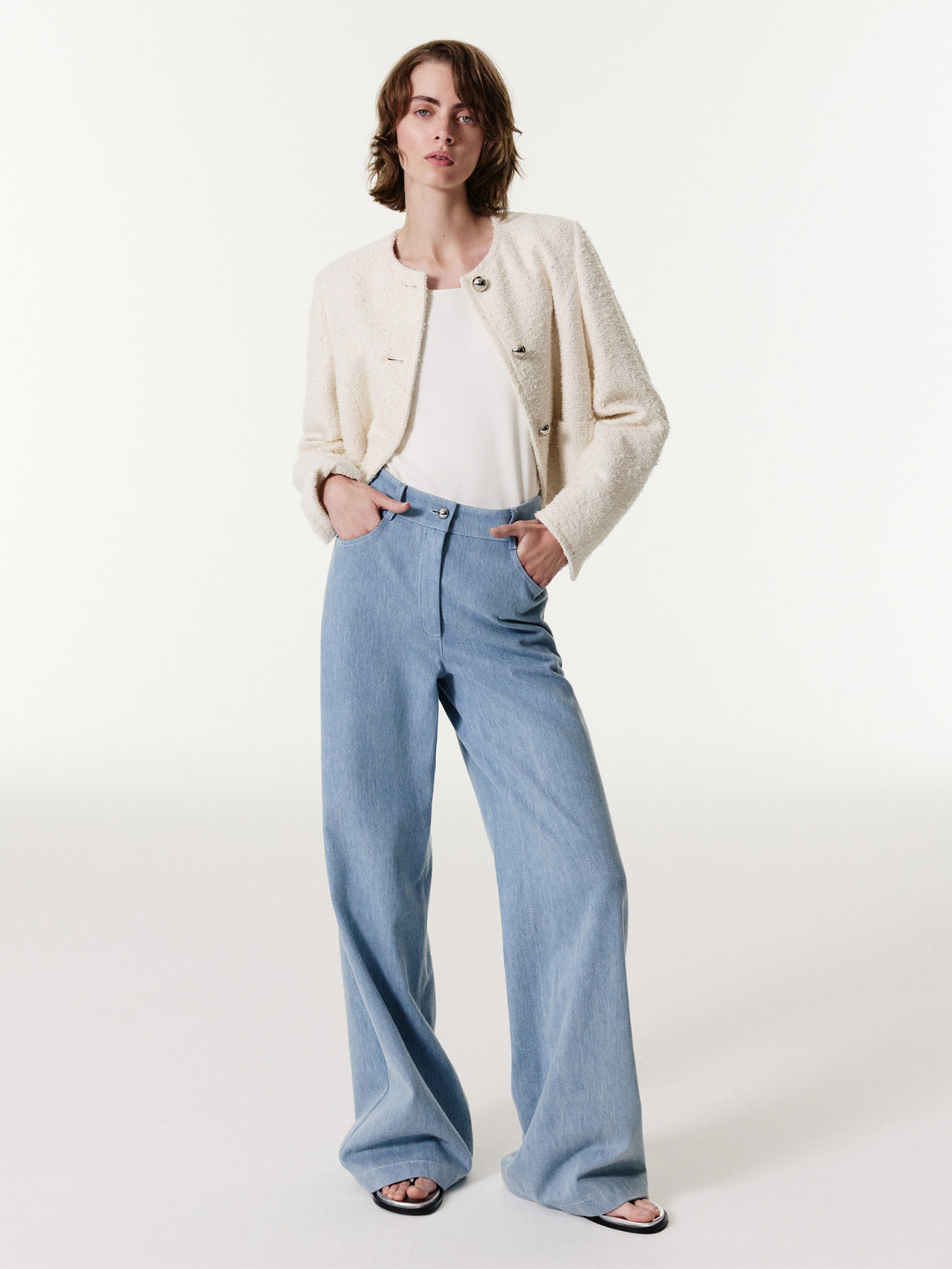Chic model in cream bouclé textured jacket and light blue wide-leg jeans, showcasing a modern ensemble with sandals against a white studio background.