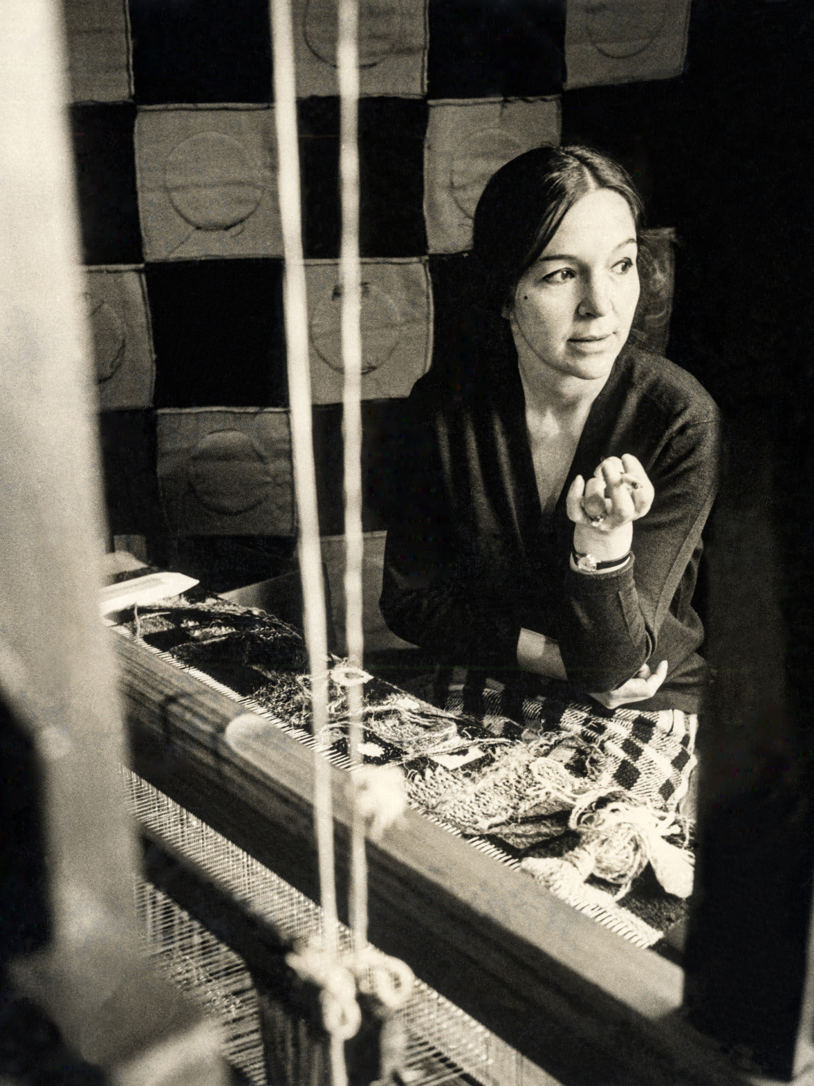 A nostalgic black-and-white portrait of artist Olga de Amaral working at her loom, capturing a moment of creative reflection amidst woven textures.