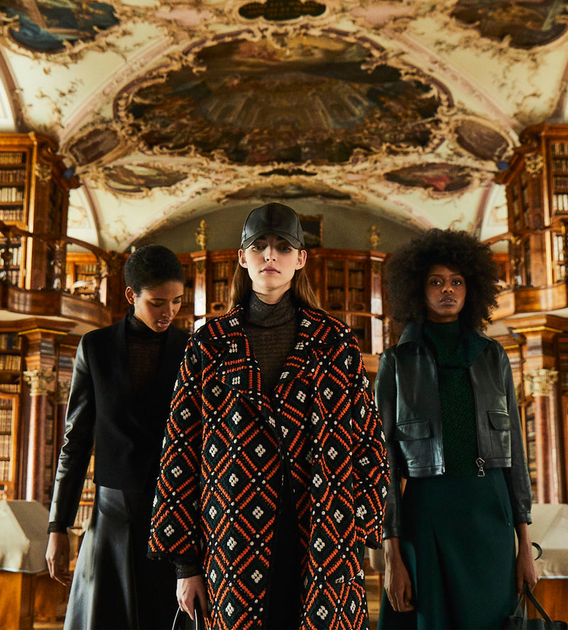Three models in the Abbey Library in St. Gallen, one model wears a multi-coloured coat of finest embroidery. In the background, the richly decorated ceiling can be admired.