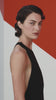 A model stands in three-quarter profile wearing a floor-length black halter gown with a deep open back. She glances over her shoulder against a minimalist gallery-like backdrop featuring bold geometric shapes in red and white.